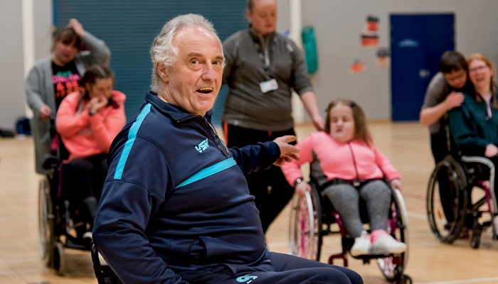 Man in a wheelchair, in a sports hall setting and pointing to a group of people in the background, 3 of the people are also in wheelchairs.