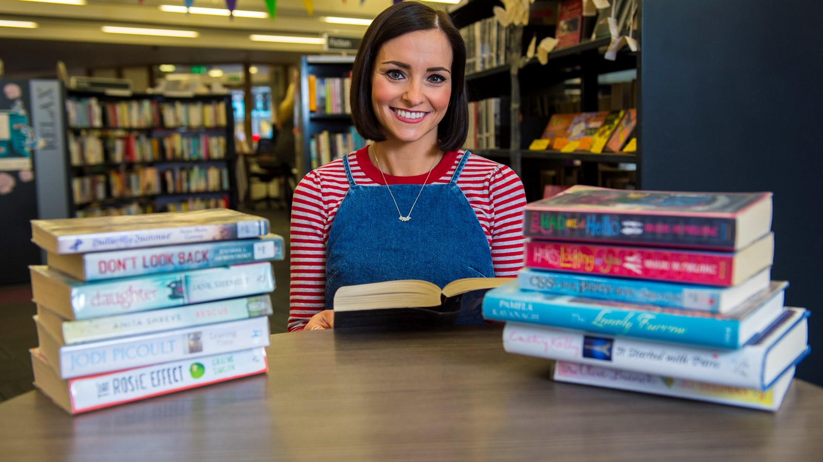 A person sitting at a table in a library between two piles of books. They are holding a book and smiling.