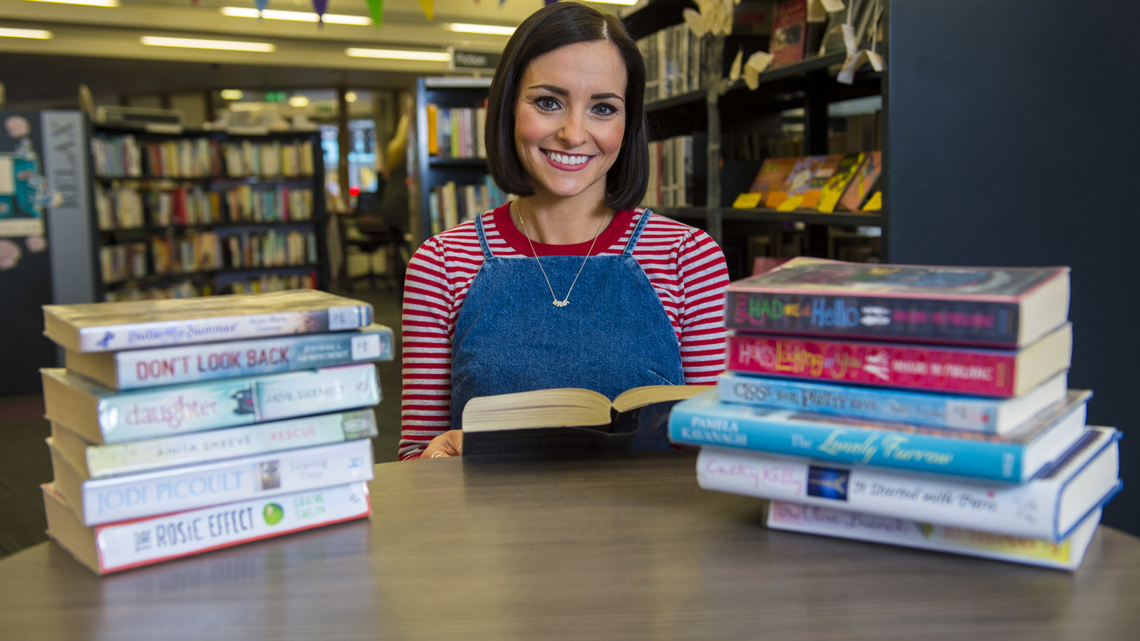 A person sitting at a table in a library between two piles of books. They are holding a book and smiling.