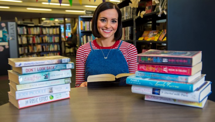 A person sitting at a table in a library between two piles of books. They are holding a book and smiling.