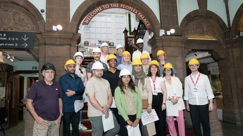 Group wearing yellow hard hats pose on stairs under a “Welcome to the Peoples Palace” sign.