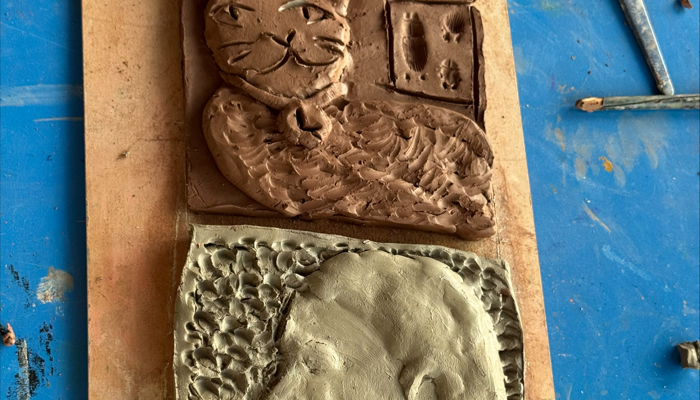 Two handmade clay reliefs displayed on a wooden board against a blue surface. The top piece, in brown clay, shows a cat with a collar lying in front of a window with two vertical panes, with detailed fur and facial features. The bottom piece, in grey clay, depicts a side profile of a person with a ponytail, surrounded by a textured background of small indentations.