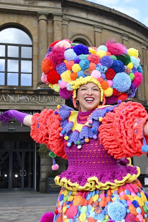 A person wearing a colourful knitted costume and a large pom pom hat, smiling with arms outstretched outside a large stone city building