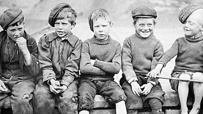 An old black and white photograph of five young people sitting on a wall. They are all wearing flat caps and their clothes are worn. Some do not have shoes on. 