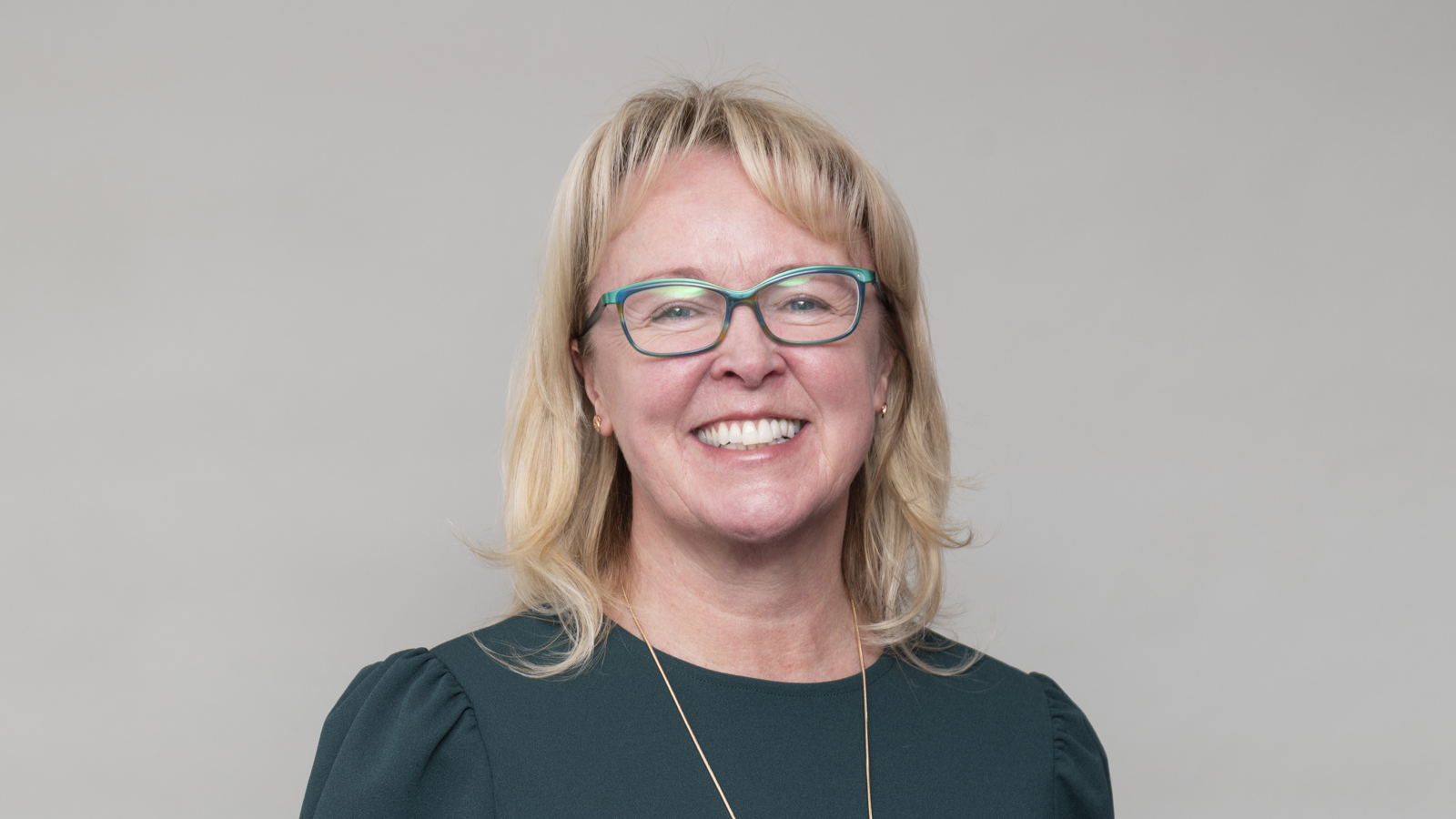 Headshot of a person with light hair and glasses, wearing a dark green top, smiling, against a neutral background.