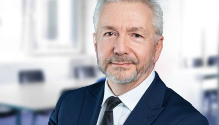 A man with grey hair dressed in a suit and tie faces the camera