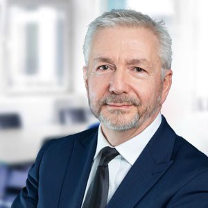 A man with grey hair dressed in a suit and tie faces the camera