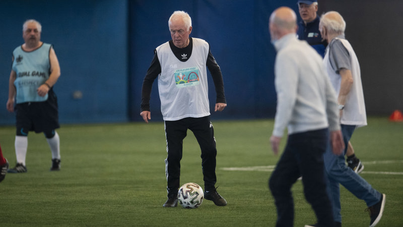 Five men on an indoor football pitch during a walking football match, one man has the ball at his feet.