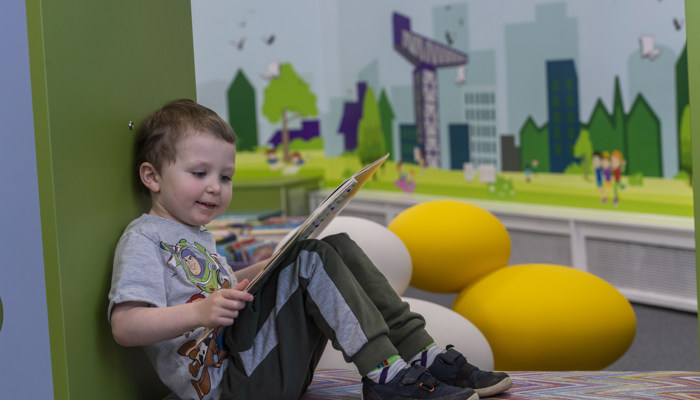 Young child sitting reading a book in Partick library. In the background is a frieze of Glasgow showing a crane, trees and people.