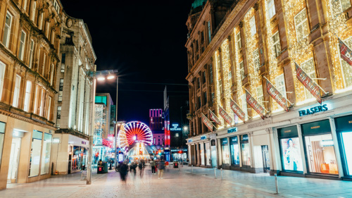 Festive Buchanan Street in Glasgow at night with illuminated shopfronts and a Ferris wheel in the distance. The Ferris wheel is located in the St Enoch's winter wonderland.