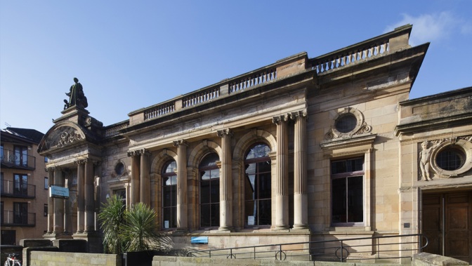 An external photograph of Woodside Library, a building made from sandstone, taken on a sunny day.