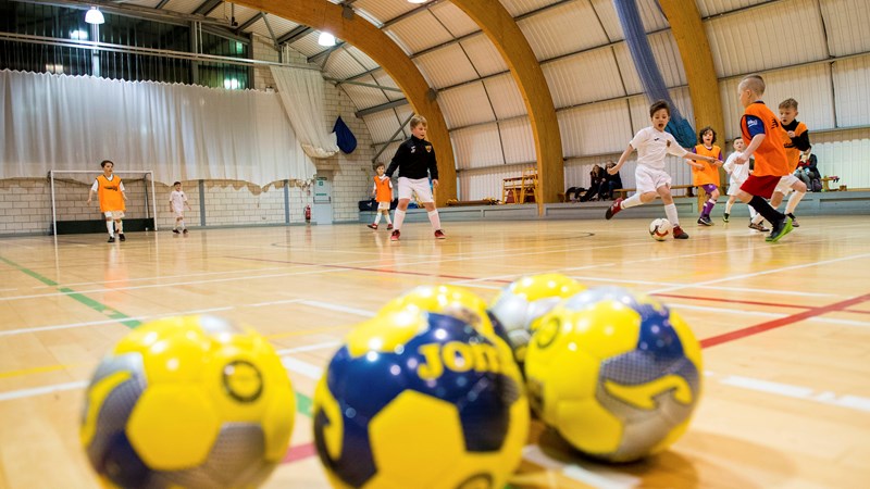 Three yellow footballs in focus in front of people playing football indoors
