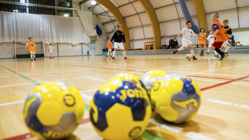Three yellow footballs in focus in front of people playing football indoors