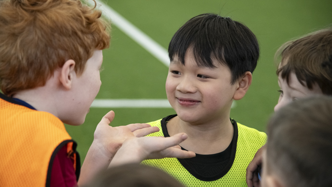 A group of young boys in sports bibs gather in conversation on an indoor turf field, with one boy smiling as another gestures animatedly.