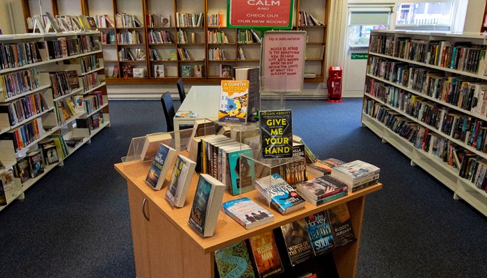 A view of the reading area of Baillieston library. There is a wooden bookstand in the middle of the room which is surrounded by more bookshelves.