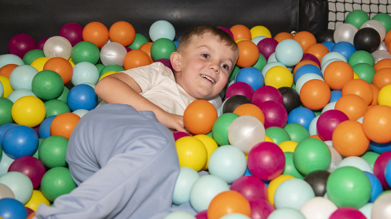 A young boy smiling while lying down in ball pit in a soft play area, the balls are a wide array of colours.