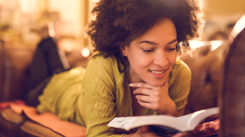 Young person smiling while relaxing and reading a book. They are lying down on their front while reading. They are wearing a mustard yellow jumper.