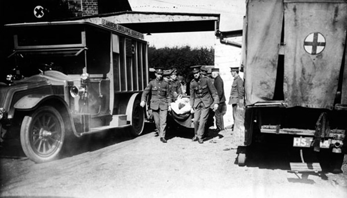 A black and white photo of several staff in uniform carrying a wounded person on a carrier to the hospital, walking between hospital transport vehicles.