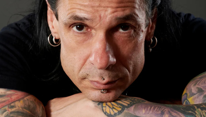 A man with long dark hair and earrings is leaning forward and resting his chin on his hands
