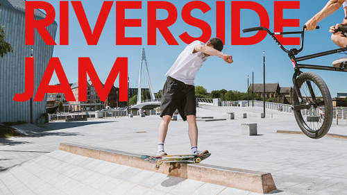 A person riding a skateboard across a concrete rail while another person performs a jump on a BMX bike. They are both on a large paved area next to a river on a clear day