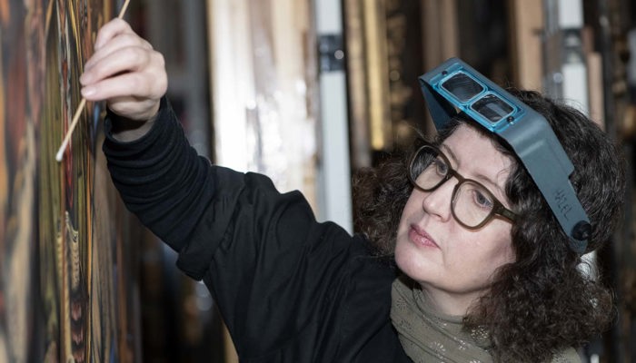Conservator wearing magnifying visor carefully cleans a painting with a small tool.