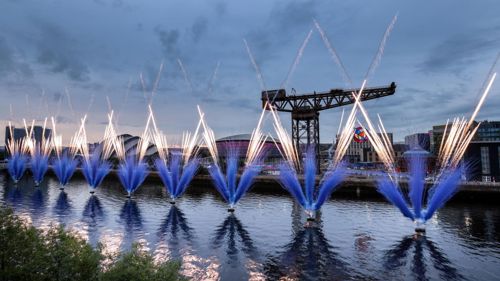 Blue and white fireworks erupt from the River Clyde at dusk, with the Finnieston Crane in the background.