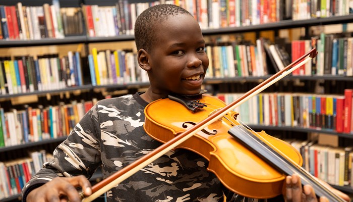 A young boy smiling while holding a violin and bow in front of shelves full of books inside The Mitchell Library in Glasgow.