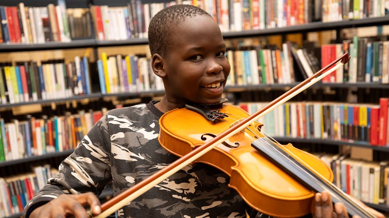 A young boy smiling while holding a violin and bow in front of shelves full of books inside The Mitchell Library in Glasgow.