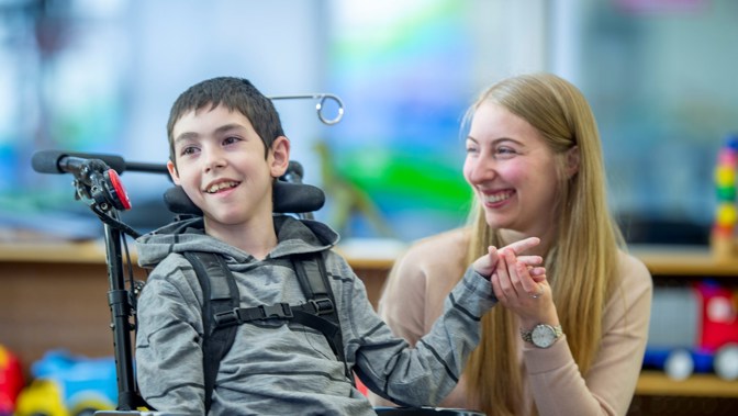 A boy in a wheelchair smiles while holding hands with a young woman who sits beside him, both laughing together in a bright indoor setting.
