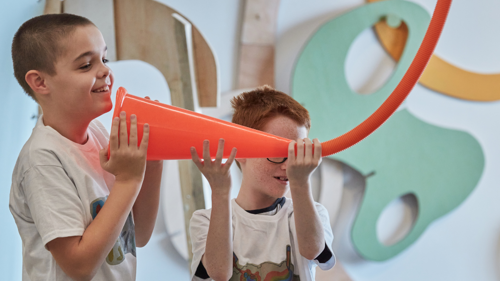 Two children playing with a bright orange cone attached to a flexible tube in a colourful, interactive exhibition.
