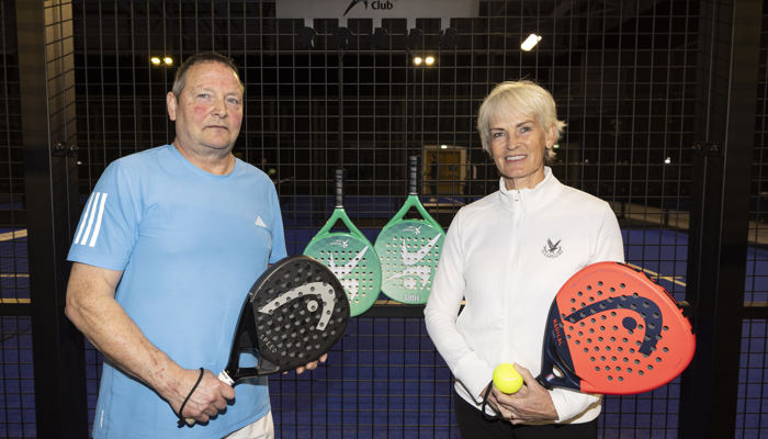 A man and a woman stand beside one another while holding padel racquets in front of jacket hooks and a sign for 'Glasgow Club'