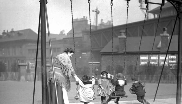 A black and white photo of a set of 3 swings in a children's playground with an adult swinging 3 young children and another young child helping.
