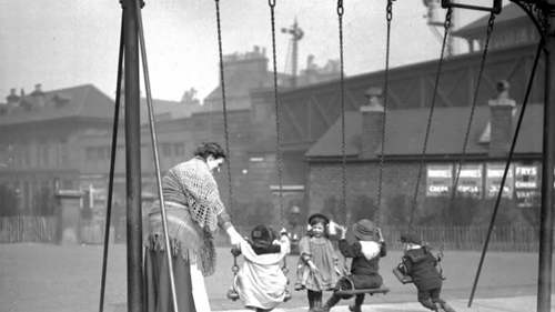A black and white photo of a set of 3 swings in a children's playground with an adult swinging 3 young children and another young child helping.