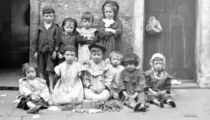 Black and white photo showing a group of 10 children of different ages in two rows, 4 children are standing in the back row whilst the rest are sitting in front row, there is a building and door behind them