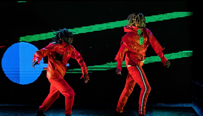 Two figures in high vis costume strike a dynamic dance pose against a dark graphic projection 