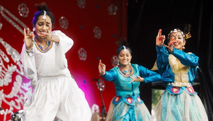 Three performers in colourful traditional costumes dance on stage with expressive hand gestures against a bright red decorative backdrop.