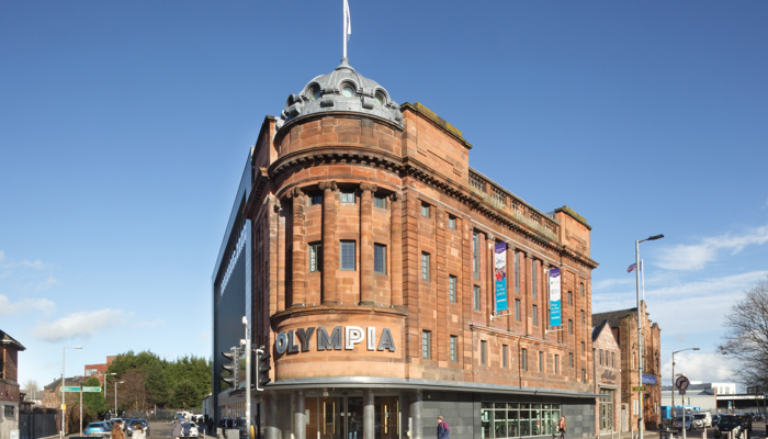 Red 4-storey sandstone corner building with a dome and a flag at the top. The sign above the doorway reads 'Olympia' in silver.