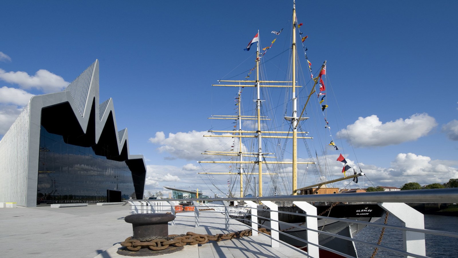 A ship with three tall masts is berthed on a river outside a museum building with a glass front
