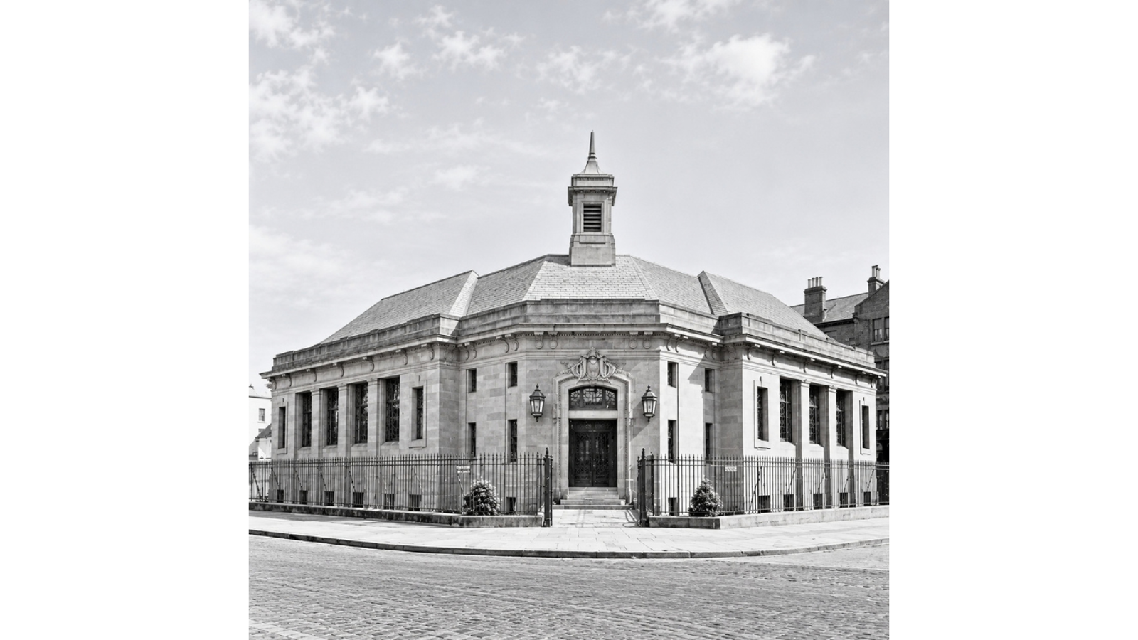 A black and white photograph of Whiteinch Library, a classic stone building on a street corner surrounded by a wrought-iron fence. The building features an ornate main entrance with double doors and decorative wall lanterns, framed by large glass windows along its outer wings and topped by a slate roof with a small central clock tower or spire.   