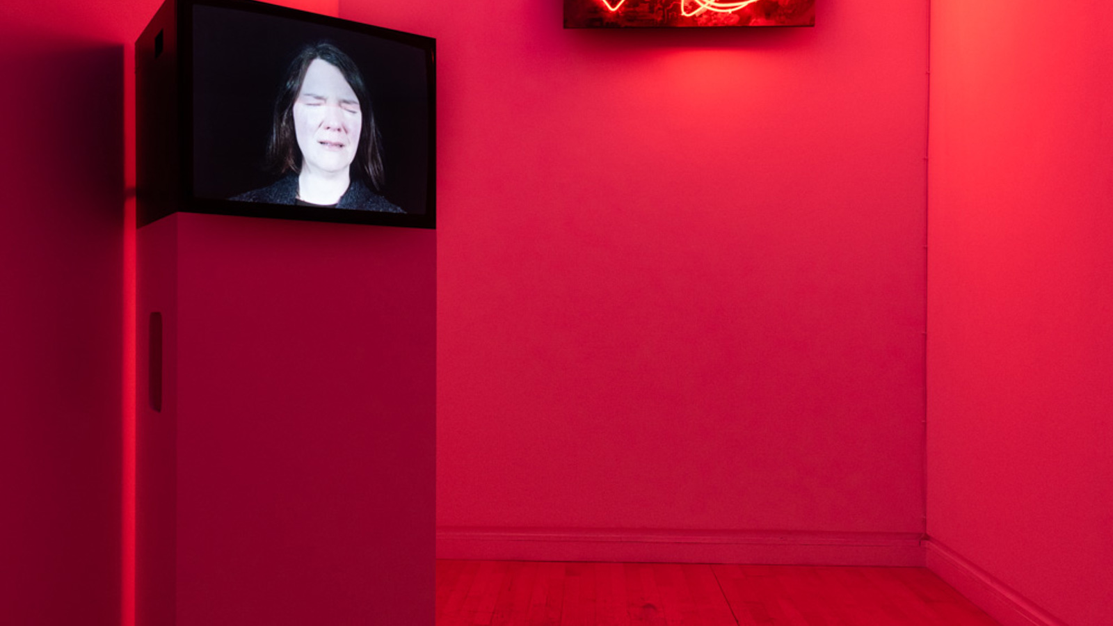 A dimly lit gallery room washed in red light features a video monitor on a white pedestal showing a close-up of a person’s face with their eyes closed. On the wall behind it, an abstract neon artwork emits shifting red patterns. The wooden floor and white walls reflect the glow, creating an immersive, atmospheric installation