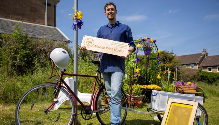 A man is pictured in front of his bike and some craft equipment. he's holding a sign that reads 'mobile print studio'