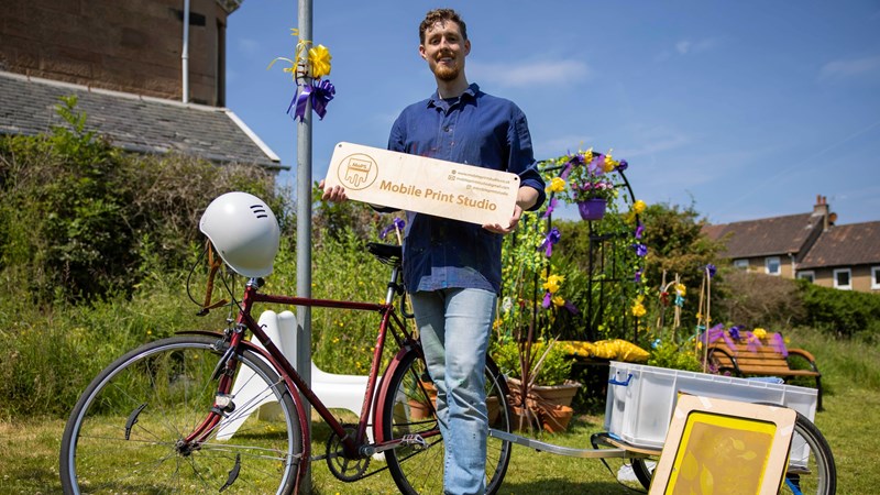A man is pictured in front of his bike and some craft equipment. he's holding a sign that reads 'mobile print studio'
