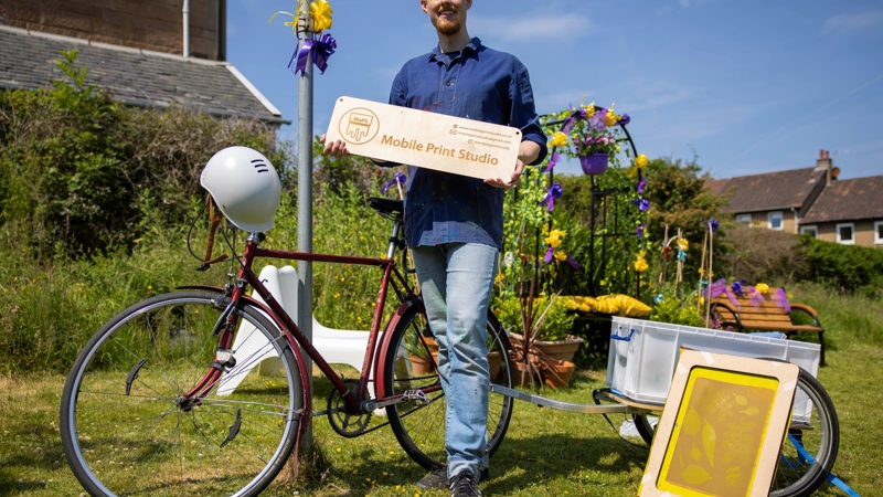 A man is pictured in front of his bike and some craft equipment. he's holding a sign that reads 'mobile print studio'