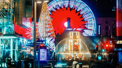 A vibrant Glasgow city centre scene featuring a large illuminated Ferris wheel with red and blue lights in the background, a glowing Christmas tree-shaped decoration in the center, and a festive carousel with bright lights in the foreground. The street is bustling with blurred figures of pedestrians, bicycles parked nearby, and colorful holiday lights adorning buildings and lampposts.