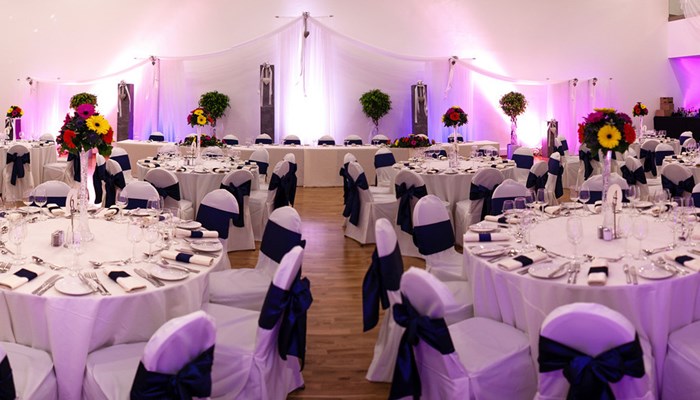 a room set up for a wedding reception. there are tables with white tablecloths, white chairs with blue bows on the back. the tables are set for a meal