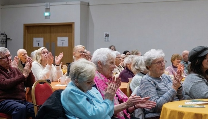 Members of the public are sitting at their tables, smiling and clapping their hands.