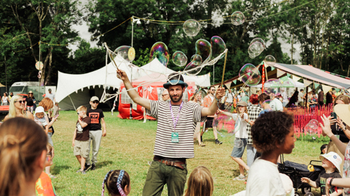 A performer is creating large bubbles in front of an audience of families at an outdoor festival on a sunny day