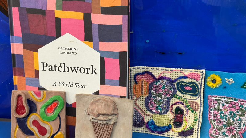 A colourful display featuring the book Patchwork: A World Tour by Catherine Legrand, shown upright with a bright patchwork-style cover. In front of it are two small clay tiles: one decorated with abstract painted shapes, the other shaped like an ice‑cream cone. To the right are two textile pieces—one on white grid fabric with embroidered flowers, and another on blue felt with appliqué floral fabric and stitched details