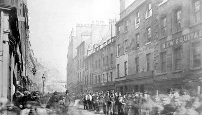 A black and white photo of a large crowd of people gathering on the pavements on a street in Glasgow, the road is cobbled and tenement buildings are on both sides.