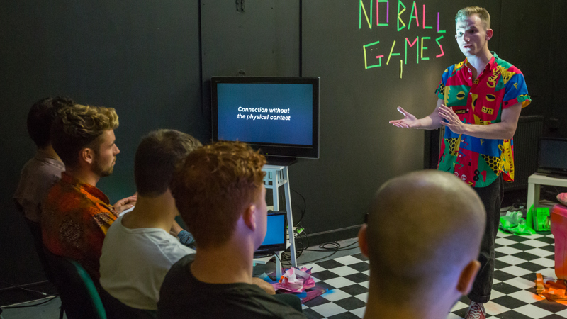 A person, is performing a play they are wearing a tropical shirt and pointing to a TV. In the background is neon tape that reads "No Ball Games".